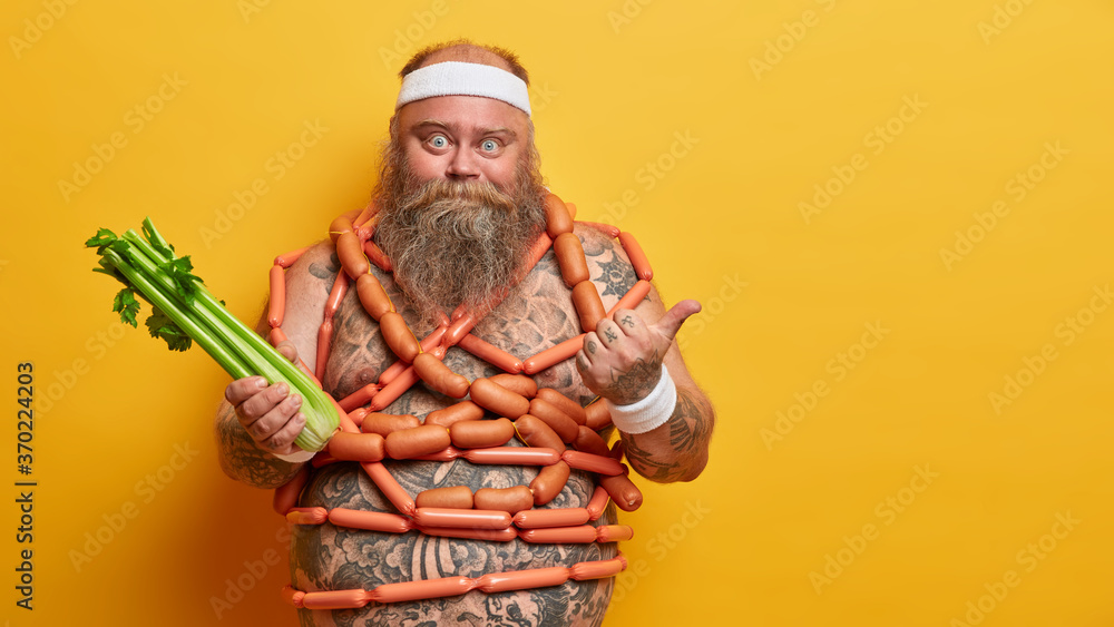Chubby man holds celery, fights against obesity, points thumb away on copy space, has temptation for eatting sausages, keeps low carb diet, forbidden to eat junk food. Nutrition, unhealthy lifestyle