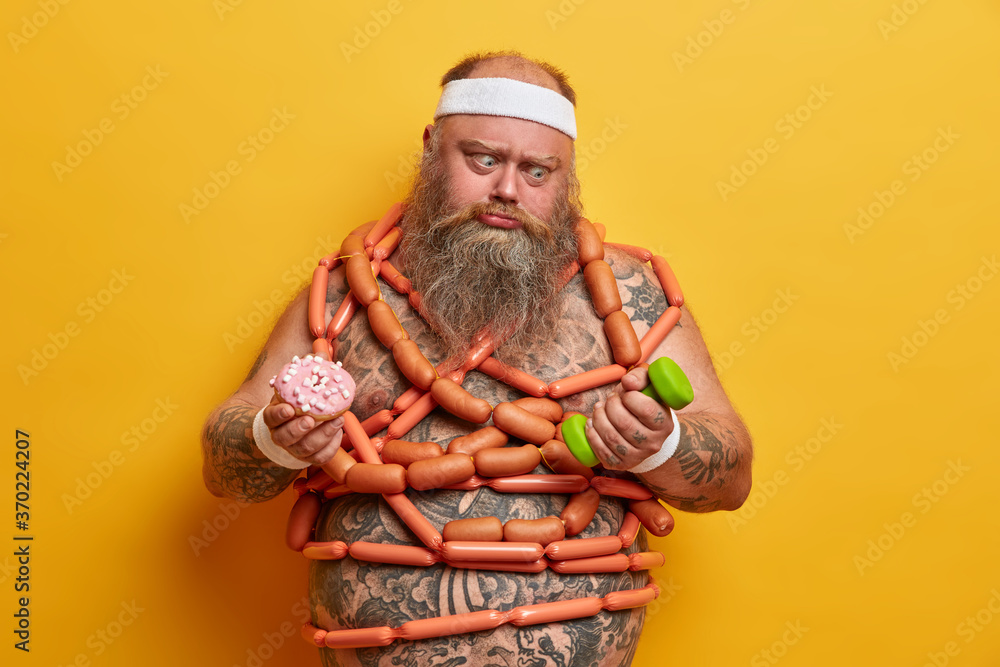 Overweight man has temptation to eat junk food goes in for sport for loosing weight has intensive cardio workout in gym holds dumbbell, doughnut cannot stop eating fast food. Body care, healthy life