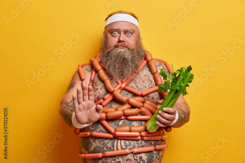Serious motivated overweight man makes stop gesture, keeps to strict vegetarian diet and chooses celery to eat, wears sport headband, stands naked, wrapped by sausages, stands indoor. Junk food