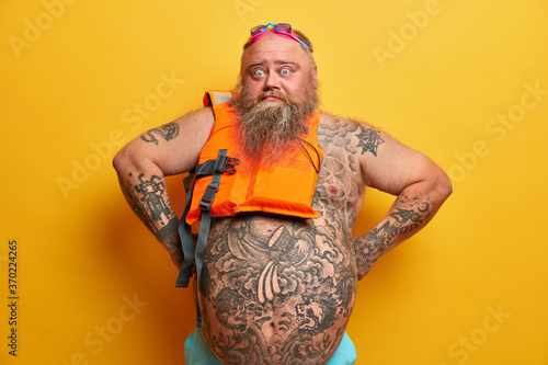 People, recreation and lifestyle concept. Surprised bearded man wears undersized swimming vest and goggles for diving poses with tattooed naked belly isolated on yellow background. Confident lifeguard
