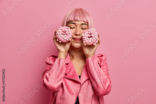 Satisfied charming woman with pink hair and fringe, closes eyes, imagines pleasant taste of donuts, dressed in rosy jacket, has sweet tooth and addiction to sugar. Monochrome indoor studio shot