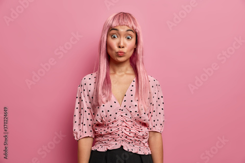 Indoor shot of surprised woman pouts lips and raises eyebrows, feels shocked to look at her new hairstyle, dyed hair in pink, wears fashionable polka dot blouse. Monochrome. Emotions, expressions
