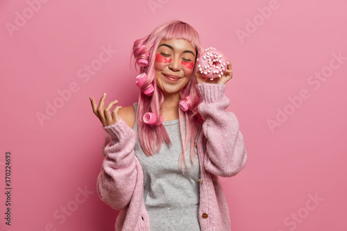 Joyful pink haired girl moves with eyes closed, holds delicious appetizing donut, wears beauty patches for skin care dressed in warm knitted sweater poses indoor rosy background. Monochrome. Junk food