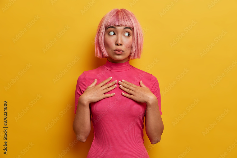 Naklejka premium Human emotions, reaction and attitude concept. Beautiful pink haired young woman keeps hands on chest and looks with wondered expression aside, has suspicion about something, feels skeptical