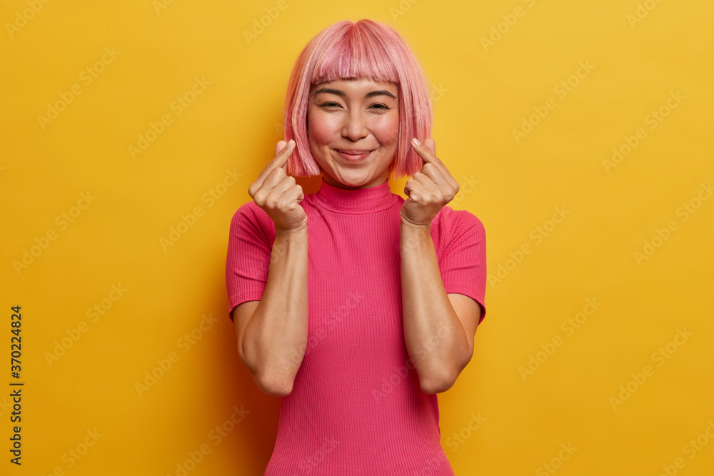 Gentle smiling pretty woman with trendy pink haircut, makes korean like gesture, expresses love, being in good mood, dressed in bright rosy t shirt, isolated on yellow wall. Body language concept. Gentle smiling pretty woman with trendy pink haircut, makes korean like gesture, expresses love, being in good mood, dressed in bright rosy t shirt, isolated on yellow wall. Body language concept.