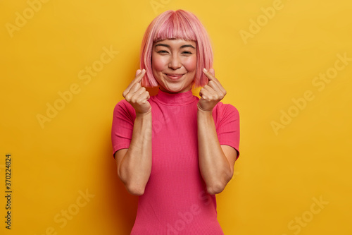 Gentle smiling pretty woman with trendy pink haircut  makes korean like gesture  expresses love  being in good mood  dressed in bright rosy t shirt  isolated on yellow wall. Body language concept.