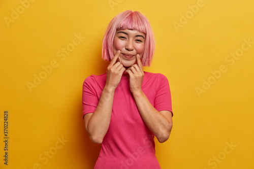 Sensual positive lady with pink hair, keeps fingers near mouth corners, forces smile, pretends having good mood, dressed in casual clothes, models against yellow background. Sincere emotions concept