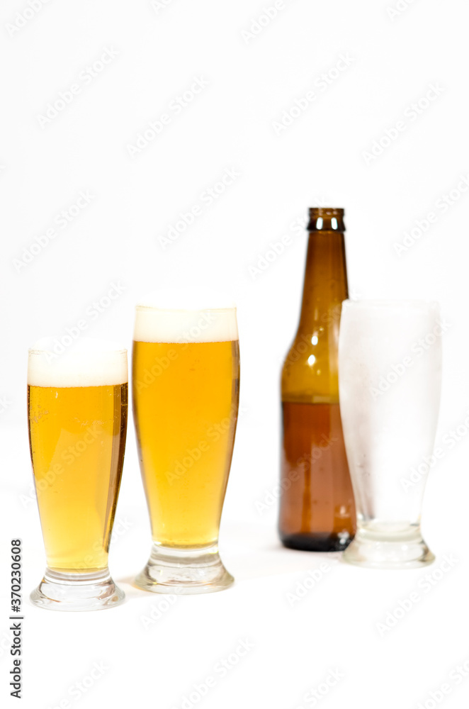 Two glasses full of beer with foam along with a brown glass bottle and another cold empty glass on a white background.