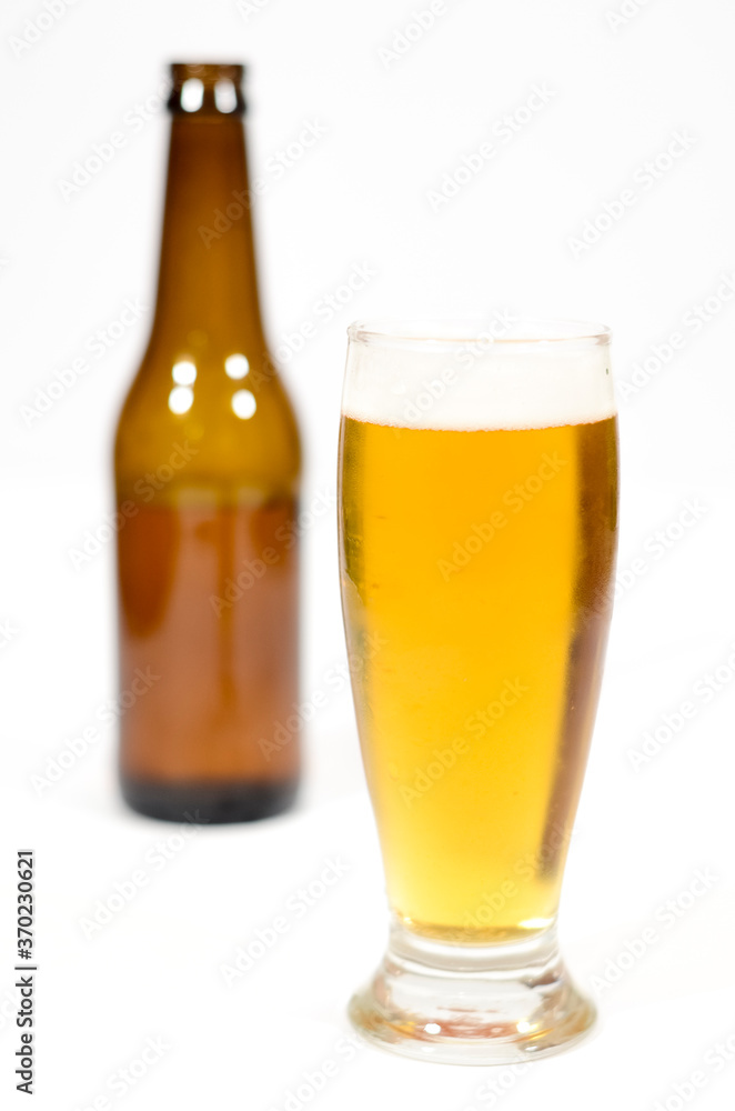 A beer glass filled with foam with a brown glass bottle to the side centered in the image with a white background.