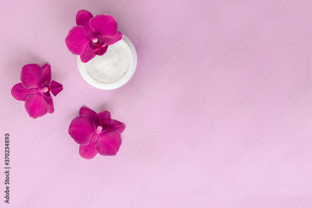 White container with cream for face and body with three magenta colored orchid flowers on pink background. Concept of delicate or eco friendly cosmetics