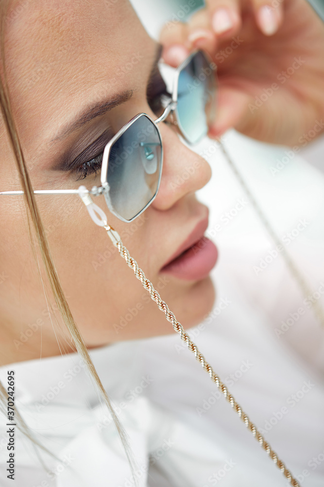 Fototapeta premium Close-up Portrait of a young beautiful fashionable woman in sunglasses. A lady poses in the open air. The model is dressed in a stylish white jacket. Women's fashion.Toned