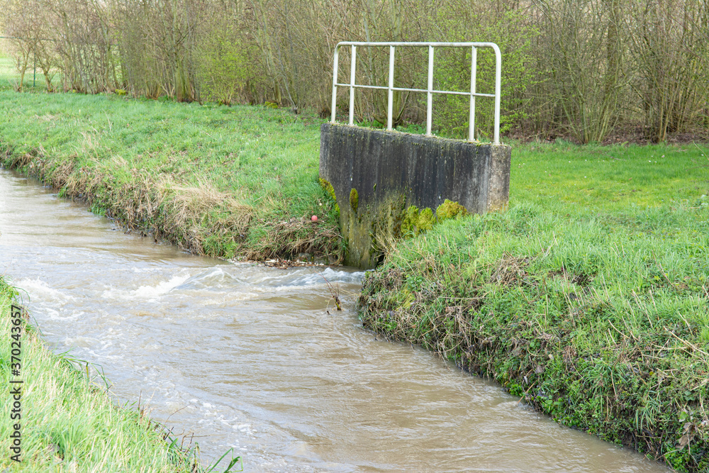 Obraz premium Close up on a small flooded river, the water is brownish, a sewer outlet, grassy banks on each sides.