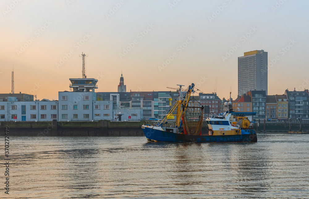 Fishing boat entering the harbor of Oostende (Ostend) city at dusk with the skyline and highest skyscraper in the background, Belgium.