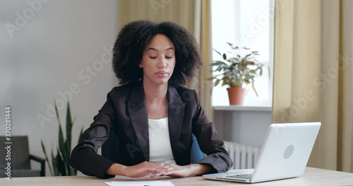 Tired, exhausted Afro girl bored of routine work study in office at home with laptop, loses energy, lays her head on her hands on table, falls asleep long day at workplace, feels sluggishly unhealthy photo