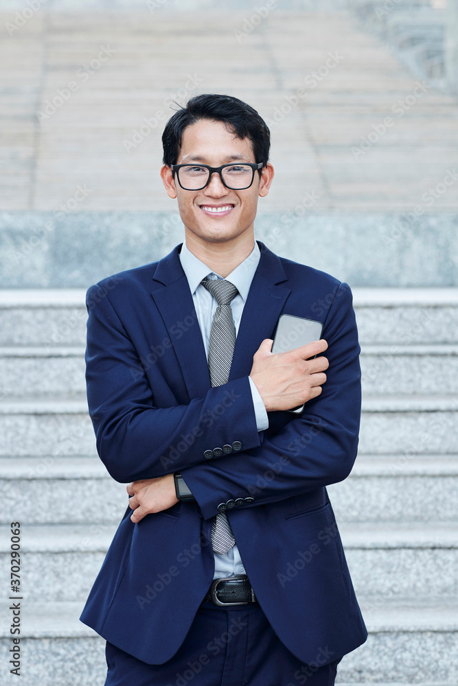 Portrait of positive young Vietnamese businessman with smartphone in hands standing outdoors and looking at camera Portrait of positive young Vietnamese businessman with smartphone in hands standing outdoors and looking at camera