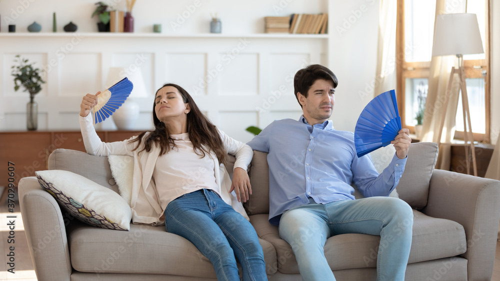Stressed tired young family couple sitting together on sofa, fanning fresh cool air with paper appliances, feeling unwell at summertime, suffering from sunstroke or high temperature at home. Stressed tired young family couple sitting together on sofa, fanning fresh cool air with paper appliances, feeling unwell at summertime, suffering from sunstroke or high temperature at home.
