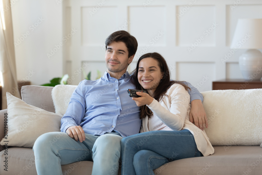 Smiling young handsome man cuddling happy beautiful wife, using remote controller, enjoying watching funny movie, comedian film or tv series, couple relaxing on comfortable couch in living room.