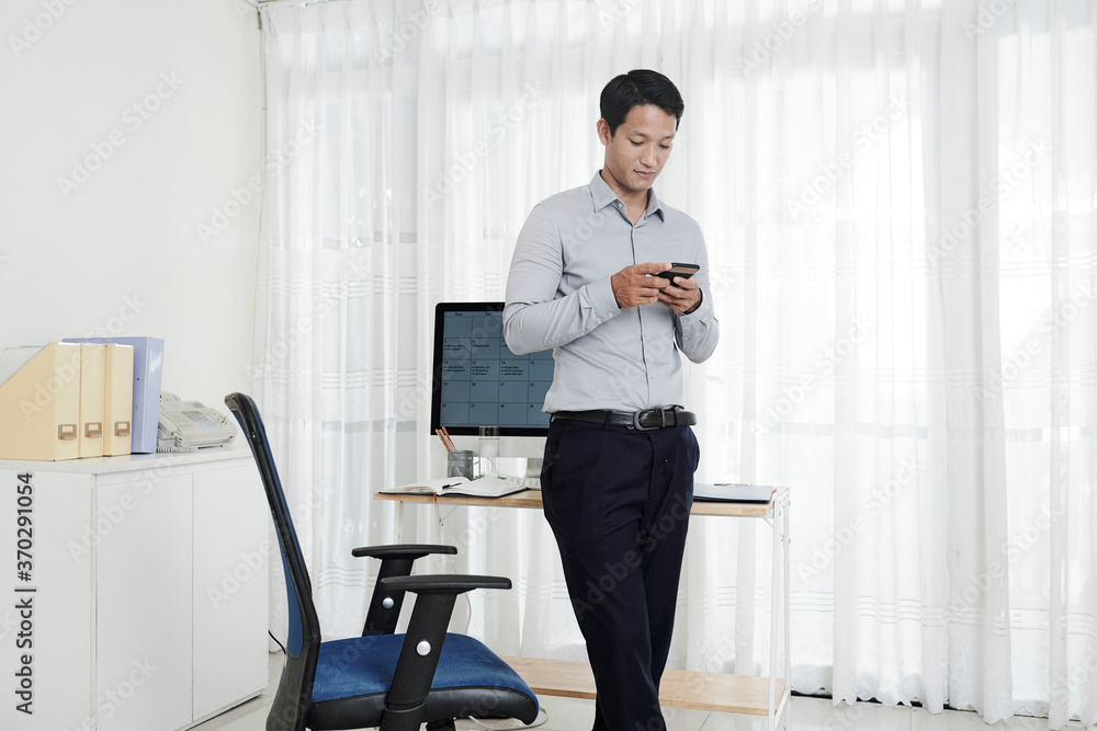 Vietnamese businessman standing in his home office and reading text message on his phone