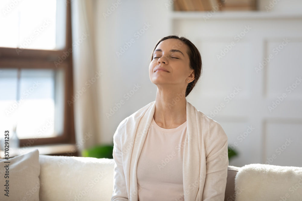Mindful young woman breathing fresh air with closed eyes, sitting on sofa alone indoors. Peaceful happy millennial girl meditating in living room, enjoying affirmations or lazy free weekend time. - obrazy, fototapety, plakaty 