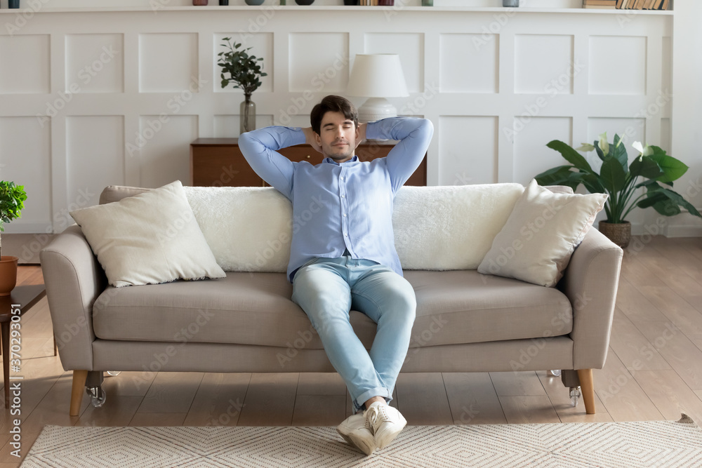 Front view peaceful young handsome man relaxing on comfortable couch, with closed eyes and folded hands behind head at home. Calm mindful millennial guy sleeping daydreaming napping in living room. Front view peaceful young handsome man relaxing on comfortable couch, with closed eyes and folded hands behind head at home. Calm mindful millennial guy sleeping daydreaming napping in living room.