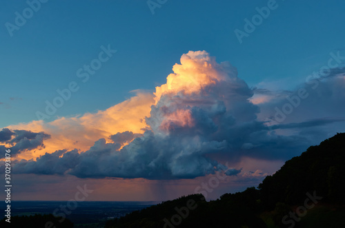 Large white weather cloud glows orange in the evening light, blue sky. trees as silouhette. Germany