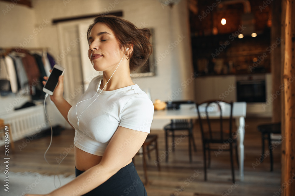Fun, relaxation, hobby and active lifestyle concept. Energetic charming young female with curvy body posing indoors in earphones closing eyes with enjoyment, dancing to music via smart phone Fun, relaxation, hobby and active lifestyle concept. Energetic charming young female with curvy body posing indoors in earphones closing eyes with enjoyment, dancing to music via smart phone