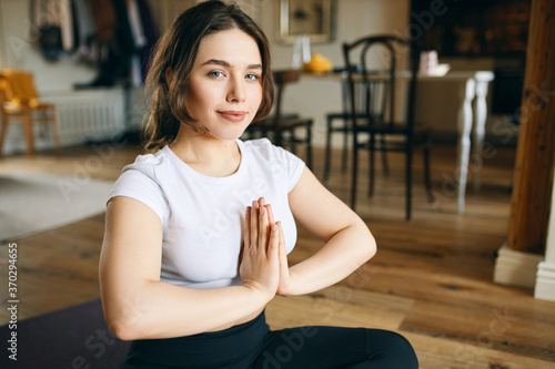 Beautiful young woman with curvy body and blue eyes sitting on floor holding hands pressed together, having refreshed relaxed facial expression after meditation, looking at camera with mindful smile