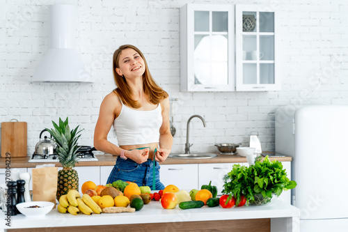Woman holds measuring tape around waist. Lot of fruits and vegetables on table. Dieting. © Vadim