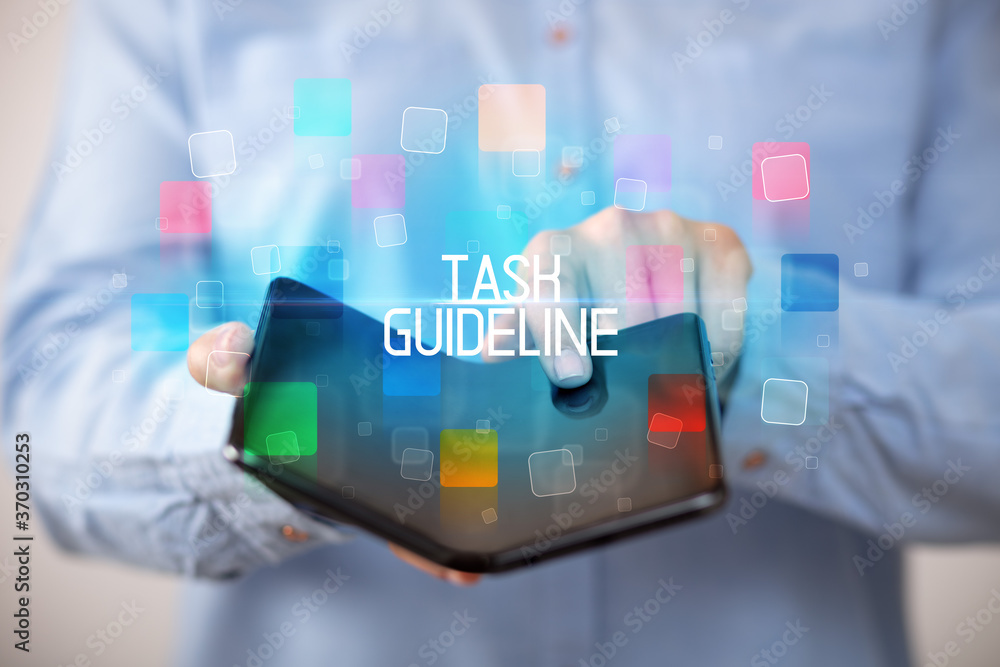 Young man holding a foldable smartphone with TASK GUIDELINE inscription, educational concept Young man holding a foldable smartphone with TASK GUIDELINE inscription, educational concept