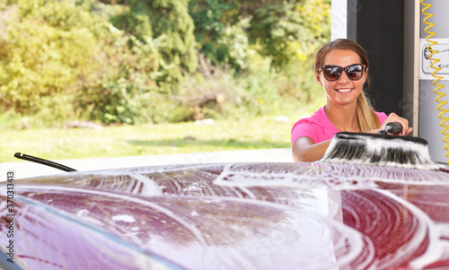 Young woman wearing pink t shirt and sunglasses cleaning her car in self serve carwash, blurred sun lit trees background