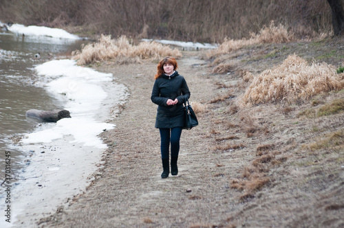Blue-eyed beautiful woman with red curly hair walks on river in winter or late autumn