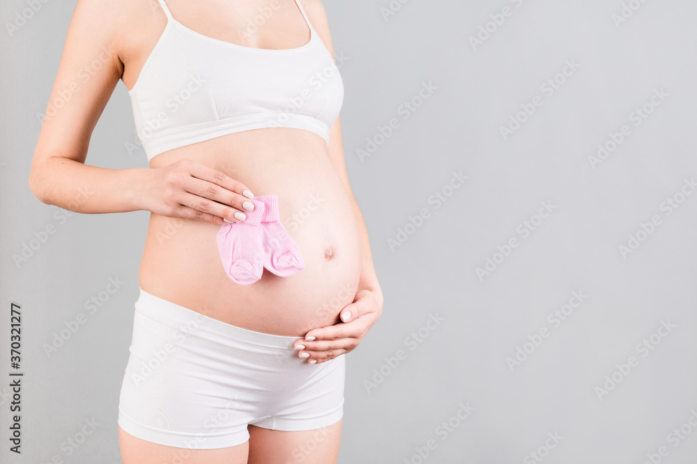 Close up portrait of pregnant woman in white underwear holding pink socks for a baby girl at gray background. Waiting for a child. Copy space Close up portrait of pregnant woman in white underwear holding pink socks for a baby girl at gray background. Waiting for a child. Copy space