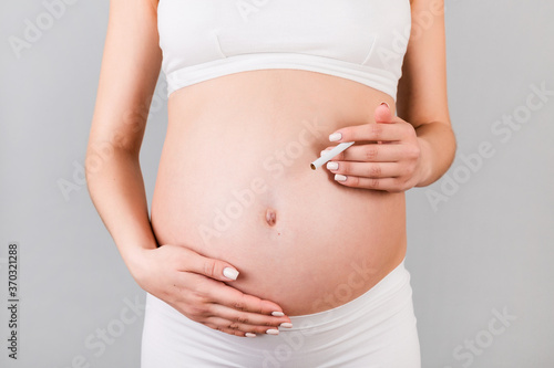 Close up of pregnant woman in white underwear with a cigarette against her belly at gray background. Dangerous risk for unborn baby