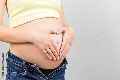 Close up of pregnant woman in unzipped jeans holding her hands in a heart shape on her belly at colorful background with copy space. Motherhood concept © sosiukin