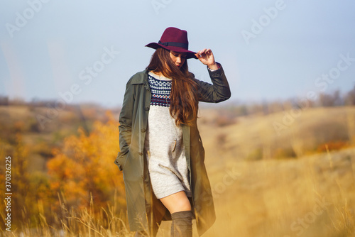 Elegant woman in coat with hat walks in autumn park. Stylish woman enjoying autumn weather in the meadow. Autumn fashion. Rest, relaxation, lifestyle concept.