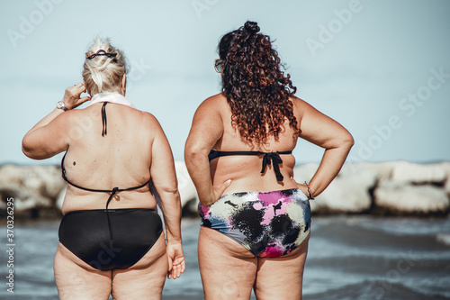 Overweight woman, mother and daughter, standing on the shore in a popular beach in Italy and looking at the sea with their arms on their hips. Vintage atmosphere. photo