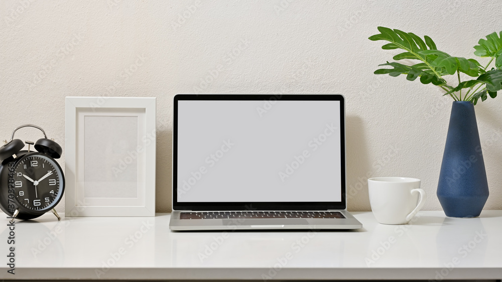 Fototapeta premium Mock up stylish of workspace with blank screen laptop, clock, frame, coffee cup and vase on white table and white background. Front view.
