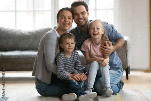 Full length portrait of happy young parents sitting on floor with joyful little preschool children siblings. Smiling couple cuddling kids, enjoying family leisure pastime together in living room.