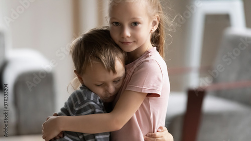 Head shot portrait of little kid girl cuddling smaller brother at home, showing love and care. Compassionate sister comforting soothing upset stressed boy in living room, siblings relations concept. photo