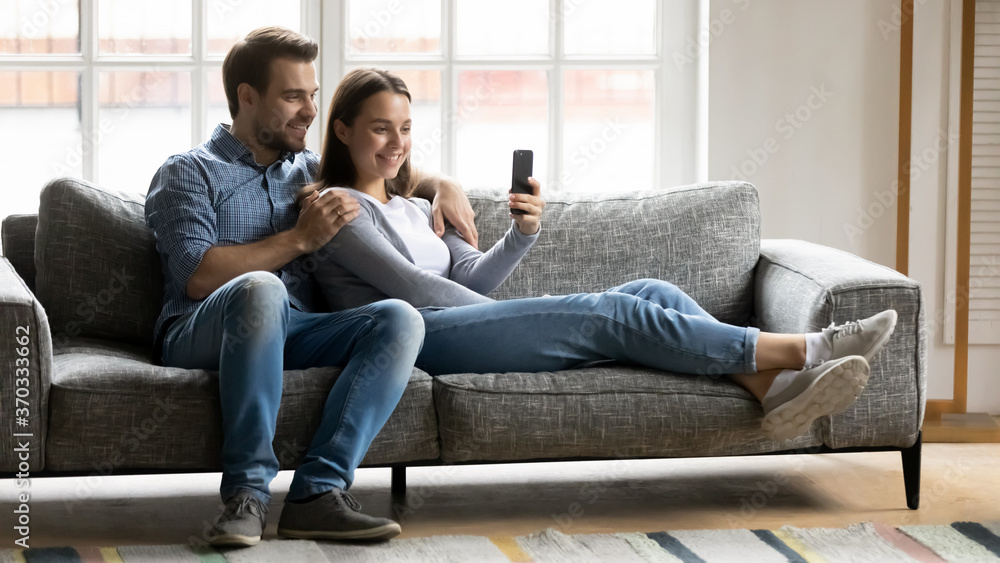 Full length relaxed young couple spending leisure time on comfortable sofa, watching funny videos in social network on smartphone, making video call or posing for selfie photo together indoors. Full length relaxed young couple spending leisure time on comfortable sofa, watching funny videos in social network on smartphone, making video call or posing for selfie photo together indoors.