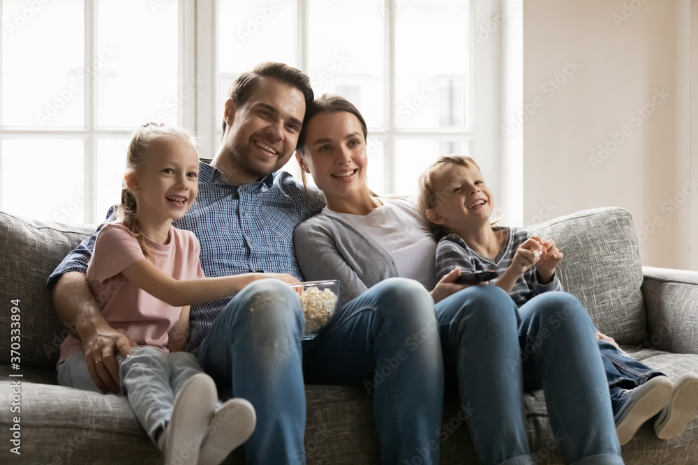 Happy bonding family of four resting on comfortable sofa, watching movie together indoors. Smiling young parents cuddling little children siblings, enjoying funny TV series with popcorn at home. Happy bonding family of four resting on comfortable sofa, watching movie together indoors. Smiling young parents cuddling little children siblings, enjoying funny TV series with popcorn at home.