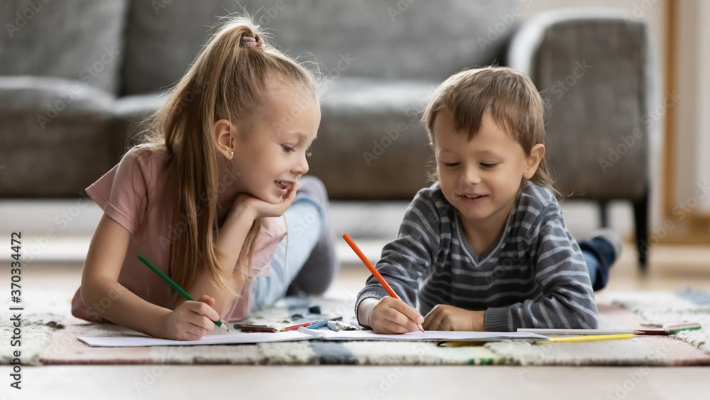 Happy cute little blonde girl lying on floor carpet with smiling funny preschool brother, enjoying drawing pictures with colorful pencils in paper albums indoors, kids weekend pastime at home. Happy cute little blonde girl lying on floor carpet with smiling funny preschool brother, enjoying drawing pictures with colorful pencils in paper albums indoors, kids weekend pastime at home.
