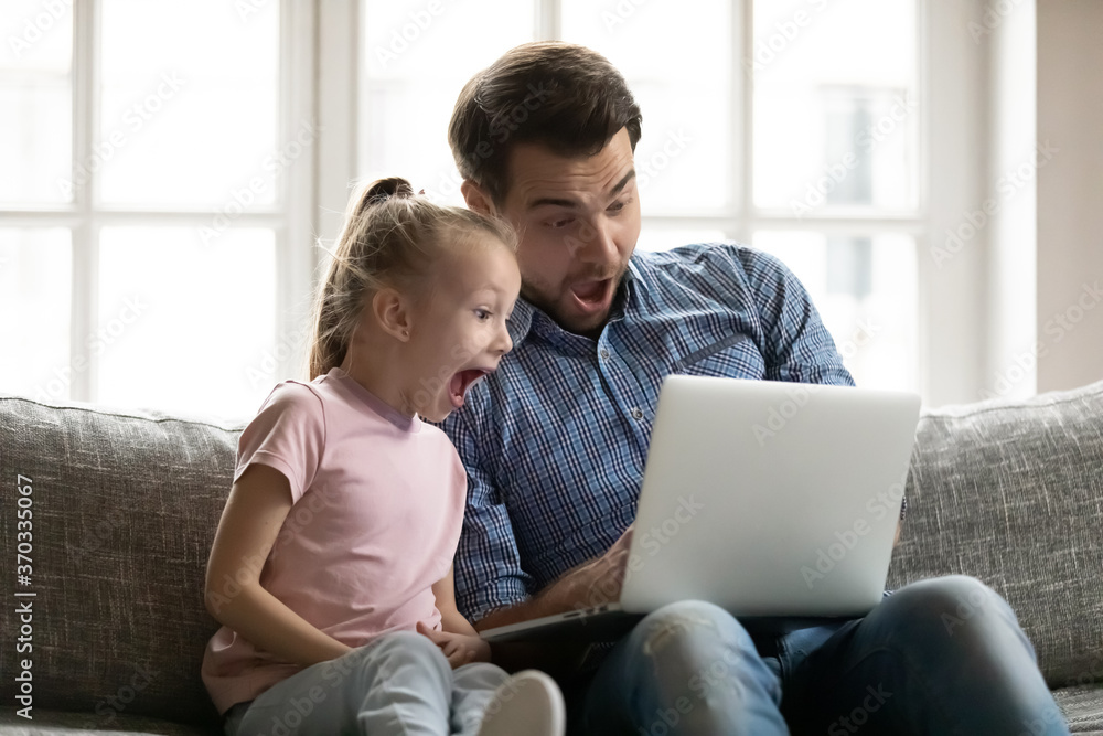 Emotional little cute child daughter watching funny movie on computer with joyful dad, sitting together on couch. Surprised two generations family looking at laptop screen, shocked by sale offer.