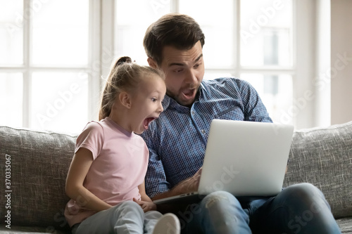 Emotional little cute child daughter watching funny movie on computer with joyful dad, sitting together on couch. Surprised two generations family looking at laptop screen, shocked by sale offer. © fizkes