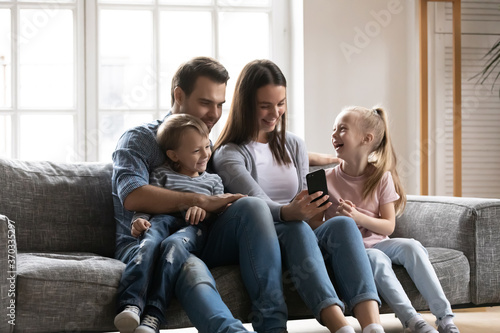 Smiling young woman showing funny video on smartphone to laughing little children and handsome father, relaxing together on comfortable sofa in living room, family weekend hobby pastime concept.