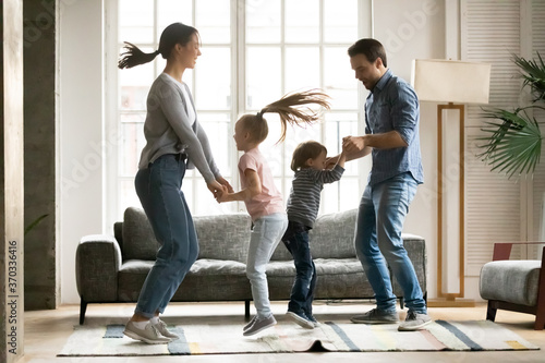 Full length positive energetic young parents holding hands of little kids siblings, jumping together in living room. Overjoyed family couple involved in dancing activity with small children at home.