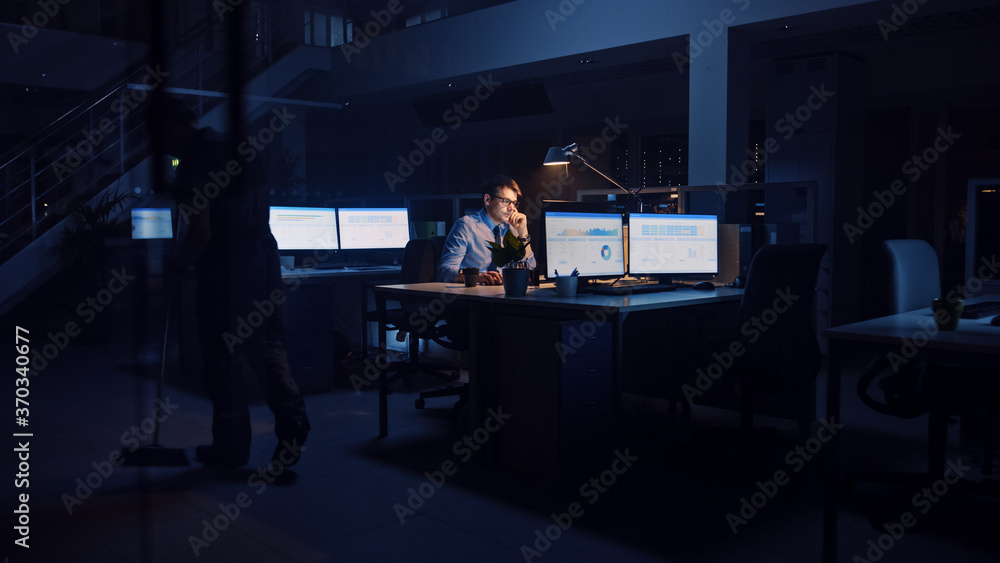 Working Late at Night in Office: Businessman Uses Desktop Computer, Analyzing, Using Documents, Solving Problems, Finishing Important Project. Ambitious Young Worker. Cleaning Man Swipes Floor Working Late at Night in Office: Businessman Uses Desktop Computer, Analyzing, Using Documents, Solving Problems, Finishing Important Project. Ambitious Young Worker. Cleaning Man Swipes Floor