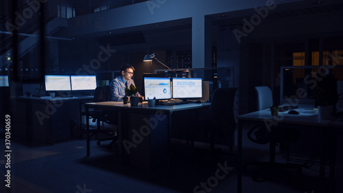 Working Late at Night in the Office: Businessman Uses Desktop Computer, Analyzing, Using Documents, Solving Problems, Finishing Important Project. Diligent Ambitious Young Worker.