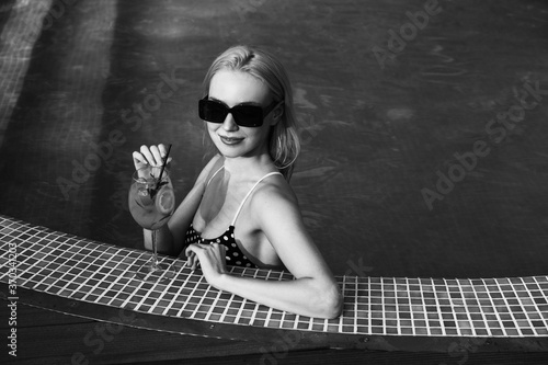 Blonde young woman in sunglasses, sitting in a pool holding a citrus cocktail. Vintage film grain picture. photo