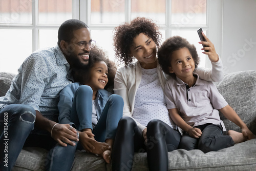 Smiling young african American family with little kids sit on sofa make self-portrait picture on smartphone together, happy biracial parents with two small children pose for selfie on cellphone